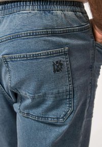 Joggers de mezclilla en azul desgastado, con cintura elástica y un bolsillo trasero con un detalle de logo bordado. Textura suave y corte relajado.