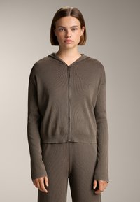 Rjava rebrasta zip-up kapuca s kapuco, dolgimi rokavi in krojeno prileganje. Material se zdi mehak in raztegljiv z gladko teksturo.