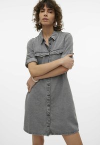 Mujer con cabello rizado que lleva un vestido de mezclilla gris con botones y mangas remangadas, cruzando los brazos y usando pendientes de aro y una pulsera de perlas.