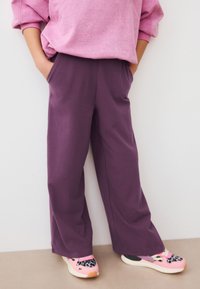 Fialové kalhoty s širokými nohavicemi a kapsami spojené s barevnými teniskami s leopardím vzorem a jasnými akcenty. Pohodlná růžová mikina nahoře.