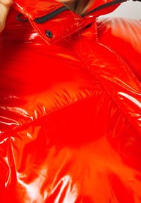 Veste rouge matelassée avec une finition brillante, dotée d'un col montant, d'une fermeture éclair et de coutures apparentes. Les accents comprennent un bouton pression et un détail de poche.