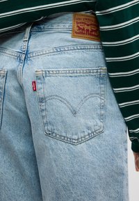 Φωτεινό γαλάζιο πίσω τσέπη τζιν Levis με κόκκινη ετικέτα και καφέ δερμάτινο σήμα μάρκας, σε συνδυασμό  με πουκάμισο με σκούρες πράσινες και λευκές ρίγες.