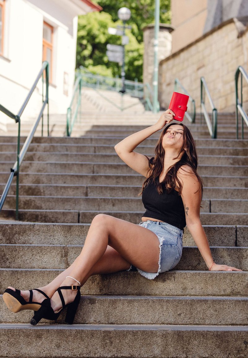 A woman sits on stone steps, wearing a black tank top and distressed denim shorts, holding a red wallet above her head, with black high-heeled shoes.
