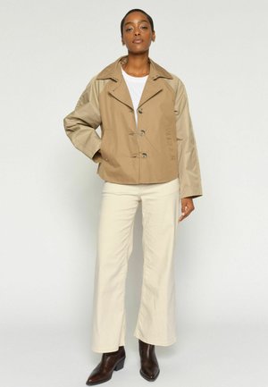 Frau steht und trägt eine beige und braune Knopfjacke, weiße Hose, weißes Hemd und dunkelbraune Stiefeletten vor einfarbigem Hintergrund.