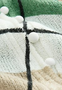 Couverture en tricot blanche, verte et beige, avec des rayures noires et des motifs texturés, ornée de trois boutons décoratifs blancs.