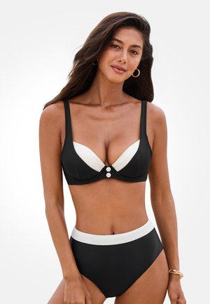 Frau mit langen dunklen Haaren trägt ein schwarz-weißes Bikini-Set mit hohem Bund und Knopfdetail, goldene Creolen und ein Armband vor weißem Hintergrund.