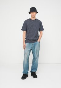 Νεαρός άντρας που φοράει μαύρο καπέλο τύπου bucket, μπλε ναυτικό ριγέ μπλουζάκι, ανοιχτό μπλε τζιν και μαύρα αθλητικά παπούτσια, στέκεται μπροστά σε ένα απλό λευκό φόντο.