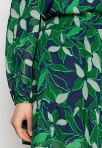 Pusero, jossa vihreä- ja valkoinen kukkakuosi tummansinisellä pohjalla. Varustettu joustavilla kalvosimilla ja kerrostetulla vyötäröllä. Kevyt kangas.