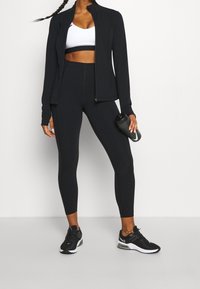 Černá sportovní bunda se zipem vpředu, ve spojení s černou sportovní podprsenkou a legínami s vysokým pasem. Černé běžecké boty a láhev na vodu jsou součástí.