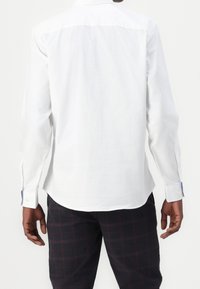 Biała koszula z długim rękawem o prostym kroju, z kołnierzykiem na guziki i akcentami na mankietach. Noszona z ciemnymi kraciastymi spodniami, widoczne detale przeszyć.