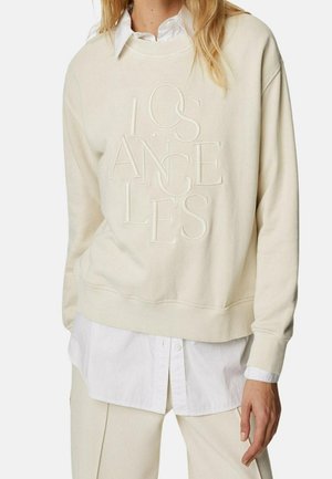 Sweatshirt couleur crème avec le texte embossé "LOS ANGELES", en coton doux, col rond et ourlet côtelé, superposé à une chemise blanche à boutons.