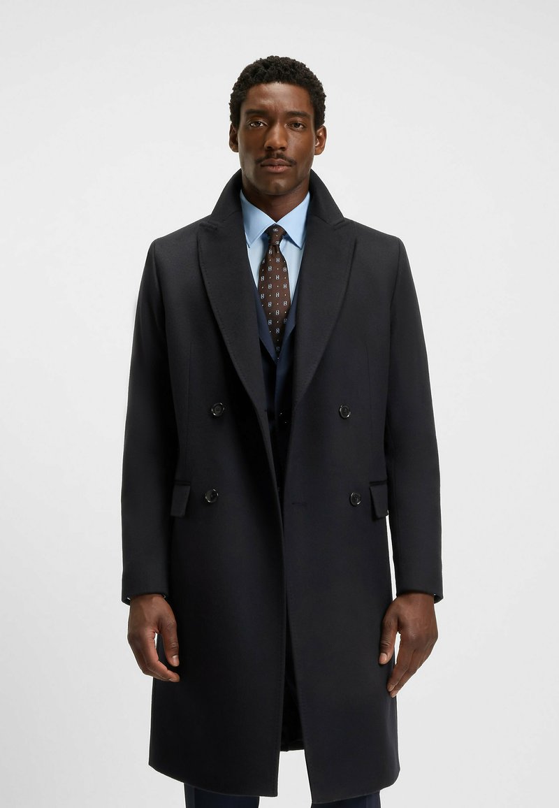 Manteau double boutonnage en mélange de laine bleu marine avec de larges revers, des accents de boutons et des poches latérales, porté sur une chemise et une cravate bleues.