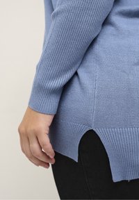 Jasnoniebieski sweter z dzianiny z prążkowanymi mankietami i dołem, z bocznym rozcięciem. Gładka tekstura, casualowy styl, zaprezentowany na czarnych spodniach.