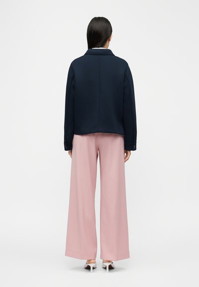 Γυναίκα που στέκεται πλάτη, φοράει μπλε σκούρο σακάκι, ανοιχτό ροζ παντελόνια φαρδιάς γραμμής και λευκά ψηλοτάκουνα παπούτσια μπροστά σε απλό φόντο.
