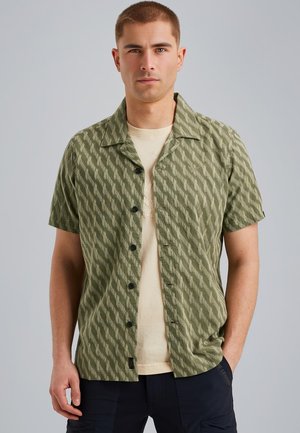Camicetta a maniche corte in verde oliva con un motivo a zigzag testurizzato, dotata di colletto e bottoni neri, indossata sopra una t-shirt beige.