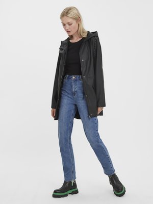 Imperméable noir et imperméable avec une capuche, porté par-dessus un haut noir, associé à un jean taille haute bleu et des bottes noires avec des semelles vertes.