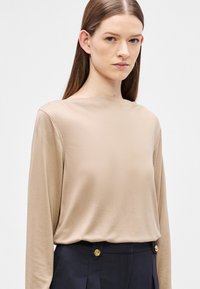 Beżowa bluzka z długim rękawem, wykonana z miękkiego materiału, z okrągłym dekoltem. Dopasowana do ciemnych spodni ozdobionych złotymi guzikami.