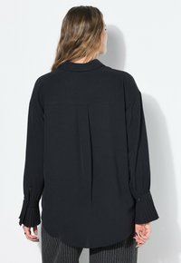 Maglietta nera a maniche lunghe con un design a polsini plissettati, dettaglio sullo yoke posteriore e orlo leggermente curvato, che mette in mostra un tessuto testurizzato.