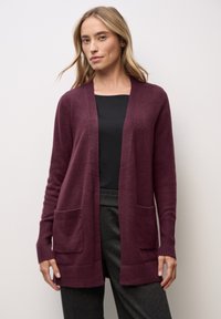 Cárdigan burdeos de corte relajado, con puños acanalados y dos bolsillos frontales. Textura suave y diseño de frente abierto. Lleva puesto sobre una blusa negra.