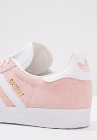 Růžové semišové tenisky Adidas Gazelle s bílými detaily, vybavené texturovanou podrážkou a zlatým nápisem "Gazelle" na boku.