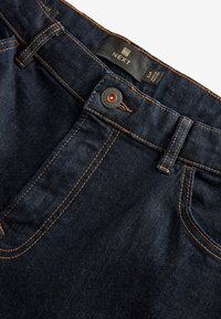 Tmavomodré džínsy z denimu s oranžovým prešitím, kovovým gombíkom, pútkami na opasok, prednými vreckami a čiernou štítkom zobrazujúcou značku a veľkosť.