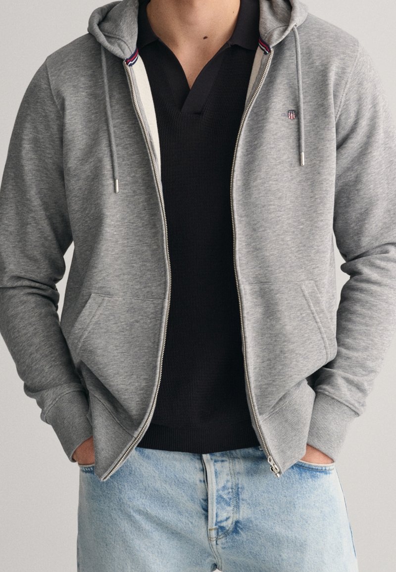 Homme portant un sweat à capuche gris clair zippé avec cordons, un t-shirt noir à col en V et un jean bleu clair, les mains dans les poches du sweat.