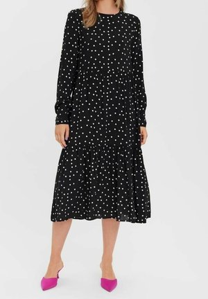 Femme portant une robe midi noire à manches longues avec des pois blancs et des escarpins pointus rose vif, debout sur un fond blanc.