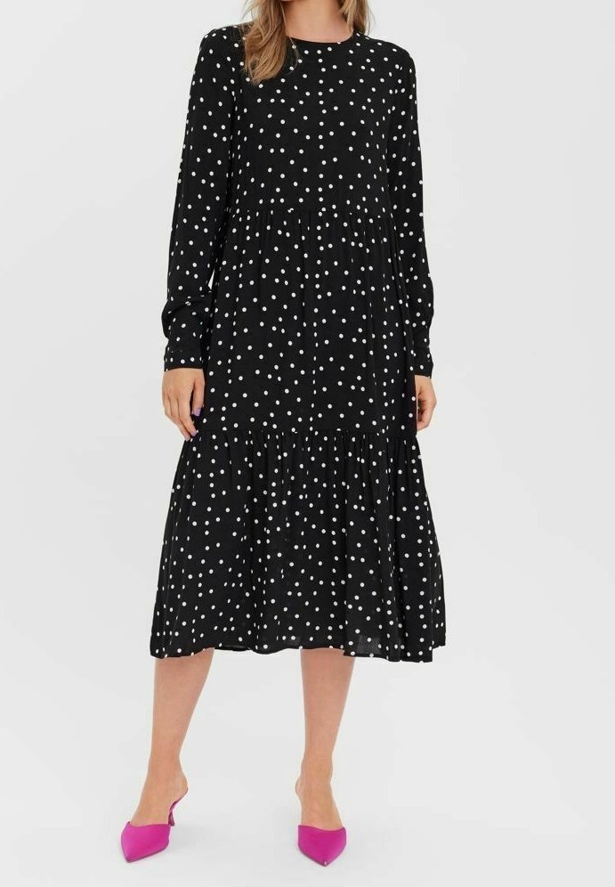 Femme portant une robe midi noire à manches longues avec des pois blancs et des escarpins pointus rose vif, debout sur un fond blanc.
