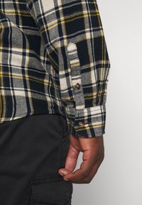 Flanellskjorta med rutigt mönster i svart, vitt och gult; har knappmanschetter och ett strukturerat tyg. Mörka byxor är delvis synliga.