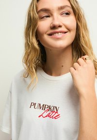 Bjela pamučna majica kratkih rukava s izvezenim natpisom "PUMPKIN Latte" u smešnim i crvenim tonovima. Ima okrugli izrez i glatku teksturu.