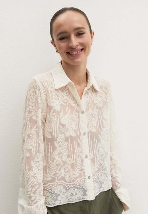 Giovane donna sorridente che indossa una camicetta bianca in pizzo floreale con bottoni e colletto e pantaloni verdi contro un muro semplice.
