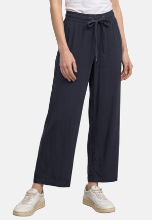 Kvinde iført marineblå vidde bukser med snøre og hvide sneakers, stående med arme afslappet ned langs siderne.