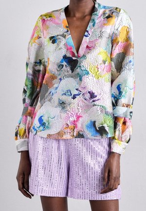 Personne portant un chemisier à manches longues avec col en V et motifs abstraits colorés, associé à un short texturé lavande.