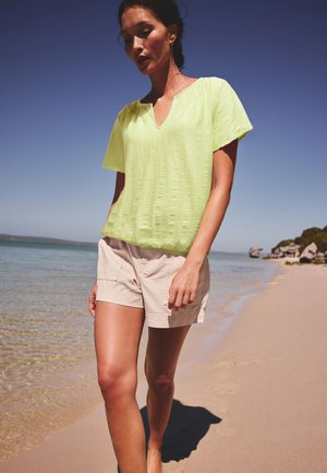 Mujer caminando descalza por una playa de arena con una camiseta de manga corta verde claro y pantalones cortos beige bajo un cielo azul claro.