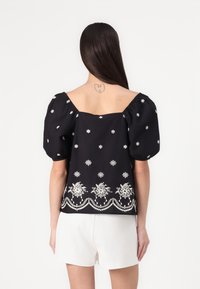 Blusa nera con maniche a sbuffo, decorata con ricami floreali bianchi e dettagli decorativi all'orlo. La parte posteriore presenta un colletto quadrato.