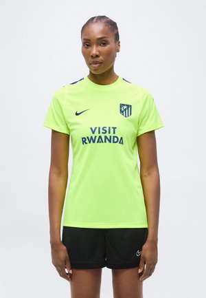 Mujer de pie con una camiseta de fútbol amarilla brillante del Atlético de Madrid con "Visit Rwanda" y pantalones cortos negros de Nike sobre un fondo liso.