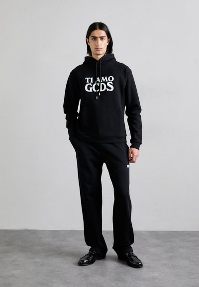 Sudadera negra con el texto "TI AMO GODS", combinada con pantalones de chándal negros. Ambas prendas están hechas de una tela suave; la sudadera cuenta con un bolsillo en la parte delantera.