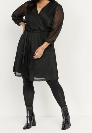 Donna che indossa un abito avvolgente nero con maniche lunghe trasparenti, collant neri e stivaletti neri lucidi, in piedi con una mano sul fianco.