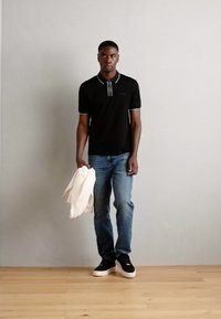 Polo noir avec col et accents blancs, associé à un jean bleu et des baskets noires, tenant une veste blanche. Contexte avec un sol en bois et un mur gris.