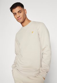 Beige sweatshirt i blødt stof med lange ærmer, rund halsudskæring og lille gult broderet logo på brystet.