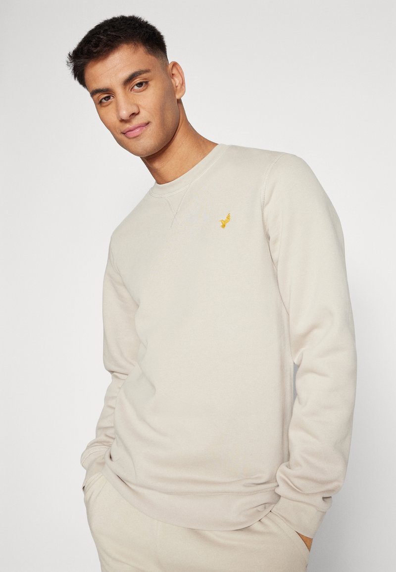 Sweatshirt beige en tissu doux avec des manches longues, encolure ronde et petit logo brodé jaune sur la poitrine.