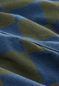 Tissu en coton présentant un motif abstrait bleu et vert olive, avec une texture douce et un tissage visible. Le design semble fluide et organique.