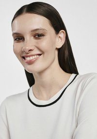 Biały sweter z czarnym prążkowanym kołnierzem, o gładkiej fakturze. Modelka ma ciemne, proste włosy i delikatny uśmiech.