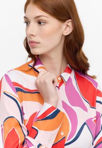 Farverig langærmet bluse med abstrakte mønstre i pink, orange og lilla. Har klassisk krave og knap-lukning ved håndleddene.