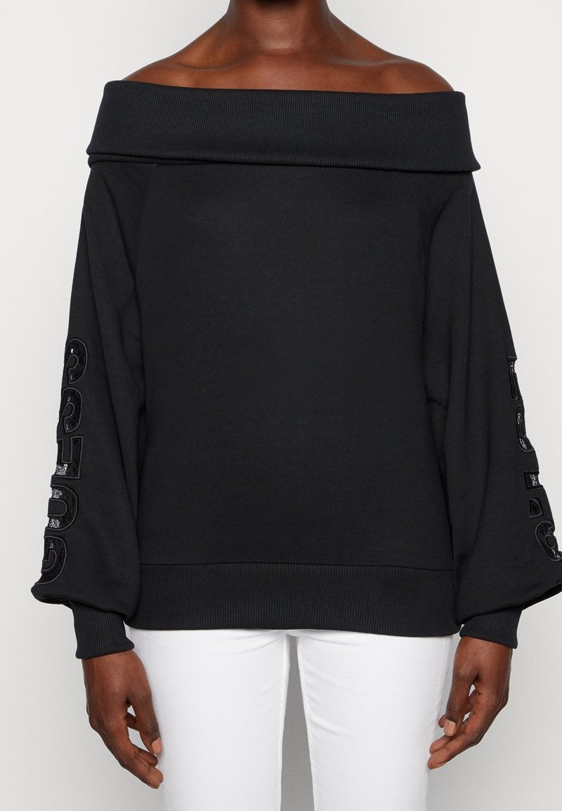 Femme portant un pull noir à épaules dénudées avec des manches bouffantes et un pantalon blanc, debout devant un fond uni.