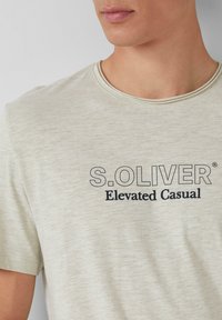 Hellbeige T-Shirt mit kurzen Ärmeln und Rundhalsausschnitt. Mit schwarzem Drucktext: "S.OLIVER" und "Elevated Casual." Weicher, strukturiert Stoff.
