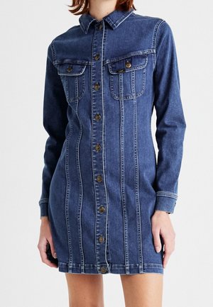 Žena v přiléhavých modrých džínových košilových šatech s knoflíkovou légou vpředu, dvěma kapsami na prsou a dlouhými rukávy na jednobarevném pozadí.