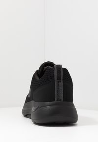 Schwarzer Sportschuh mit einem strukturierten Mesh-Obermaterial, gepolstertem Kragen und einer glatten Gummisohle. Die Rückseite hat ein vertikales Logodesign.