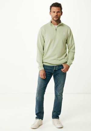 Sudadera de cremallera verde claro con cuello alto, combinada con jeans azules y zapatillas blancas. Textura suave con un detalle de logo minimalista.