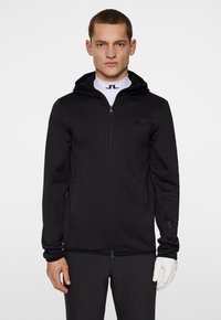 Svart zip-up jacka med huva, tillverkad av slät tyg. Har en logotyp på bröstet och en justerbar nederkant. Modellen bär ett vitt baslager.